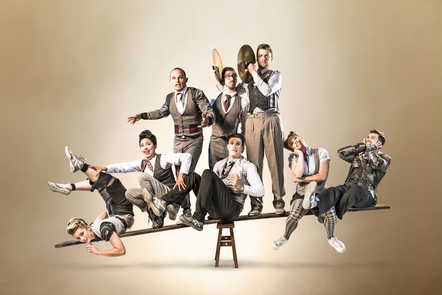 Eight circus performers in old fashioned suits balance on a plank of wood balanced precariously on a timber stool.