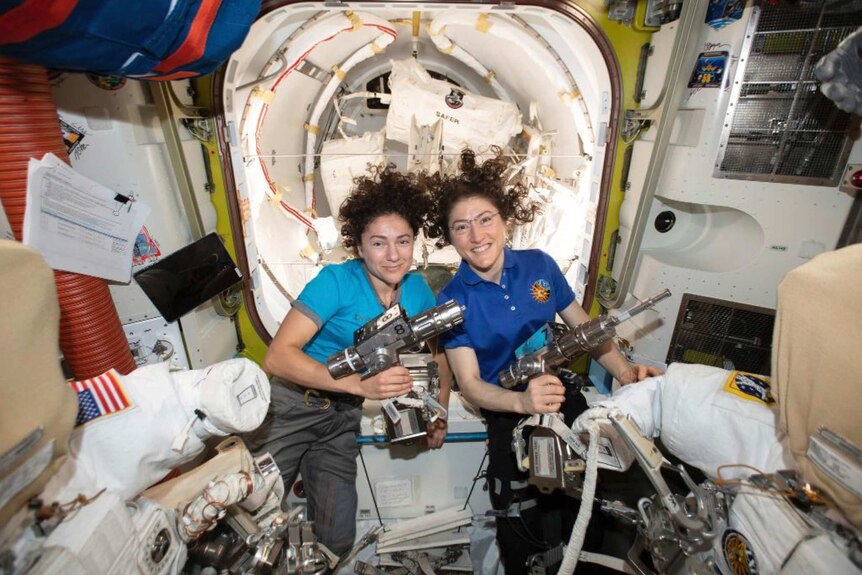 Jessica Meir, left, and Christina Koch hold silver objects as they float in the International Space Station. They both wear blue