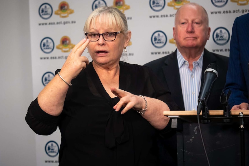 A female Auslan interpreter signs emergency information to a camera in front of an ACT Government banner.