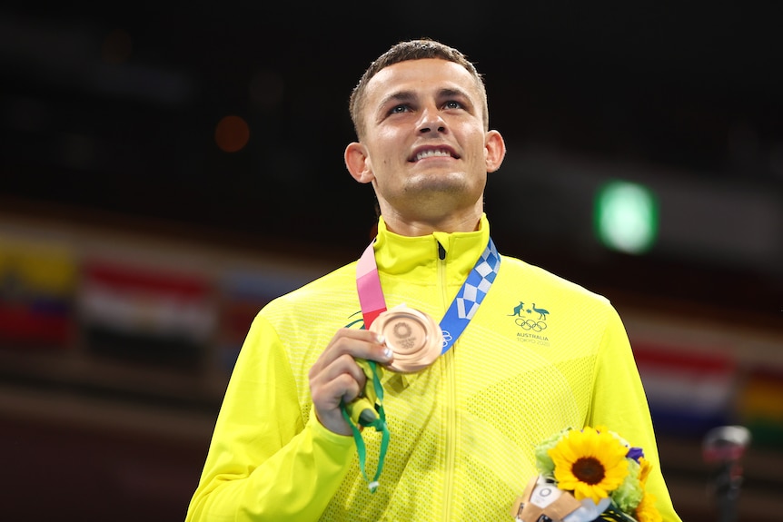 An Australian boxer stands on the podium smiling and looking up while holding his Olympic bronze medal in his hand.