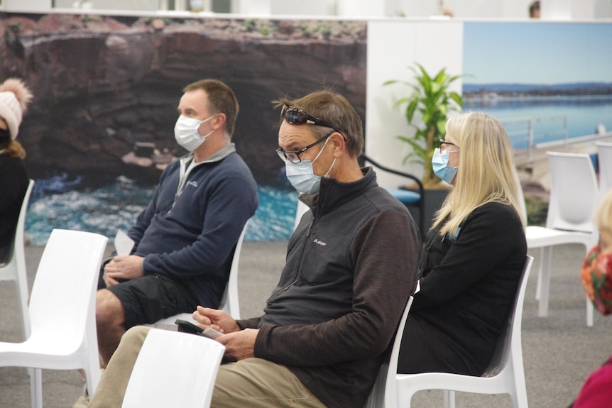 Three people wearing masks sitting on white plastic chairs.