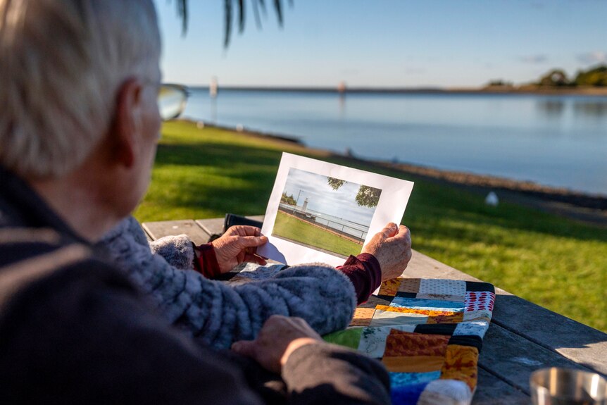 An elderly couple holds a piece of paper with a photo of the ocean up against the same coast to compare in real life.