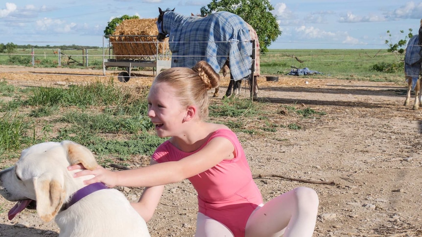 A young girl in a ballet outfit crouches with a Labrador pup, with horses behind her.