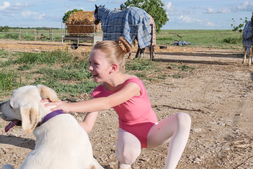 A young girl in a ballet outfit crouches with a Labrador pup, with horses behind her.