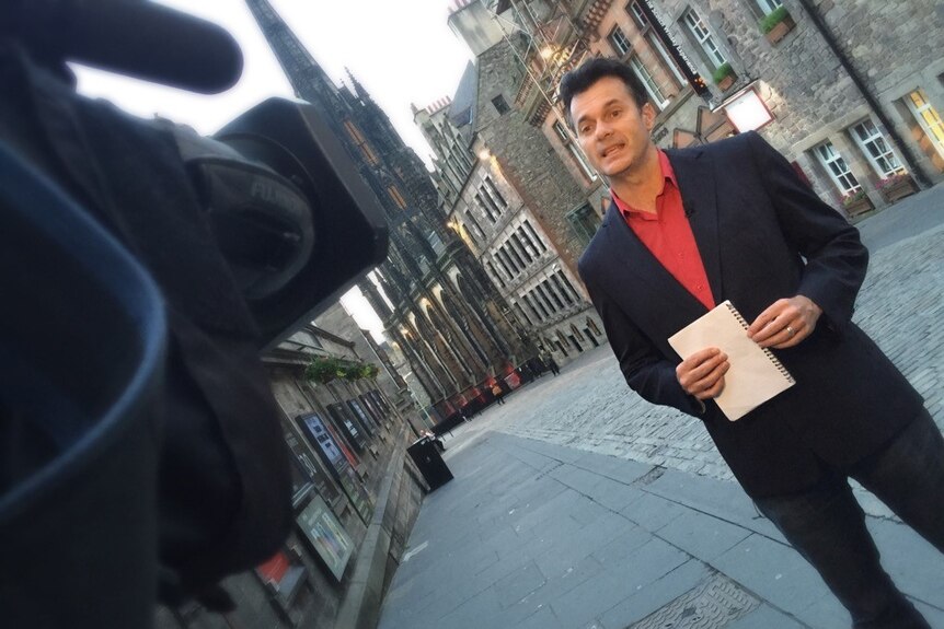 Steve Cannane holding notebook talking to camera with old stone buildings in background.