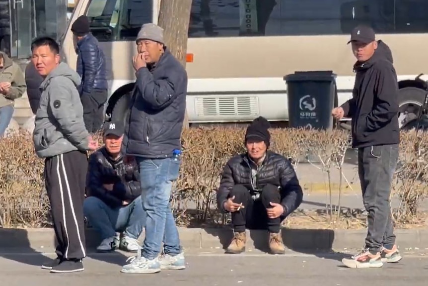 Three men stand wearing puffer jackets while a man sits next to them on a step.