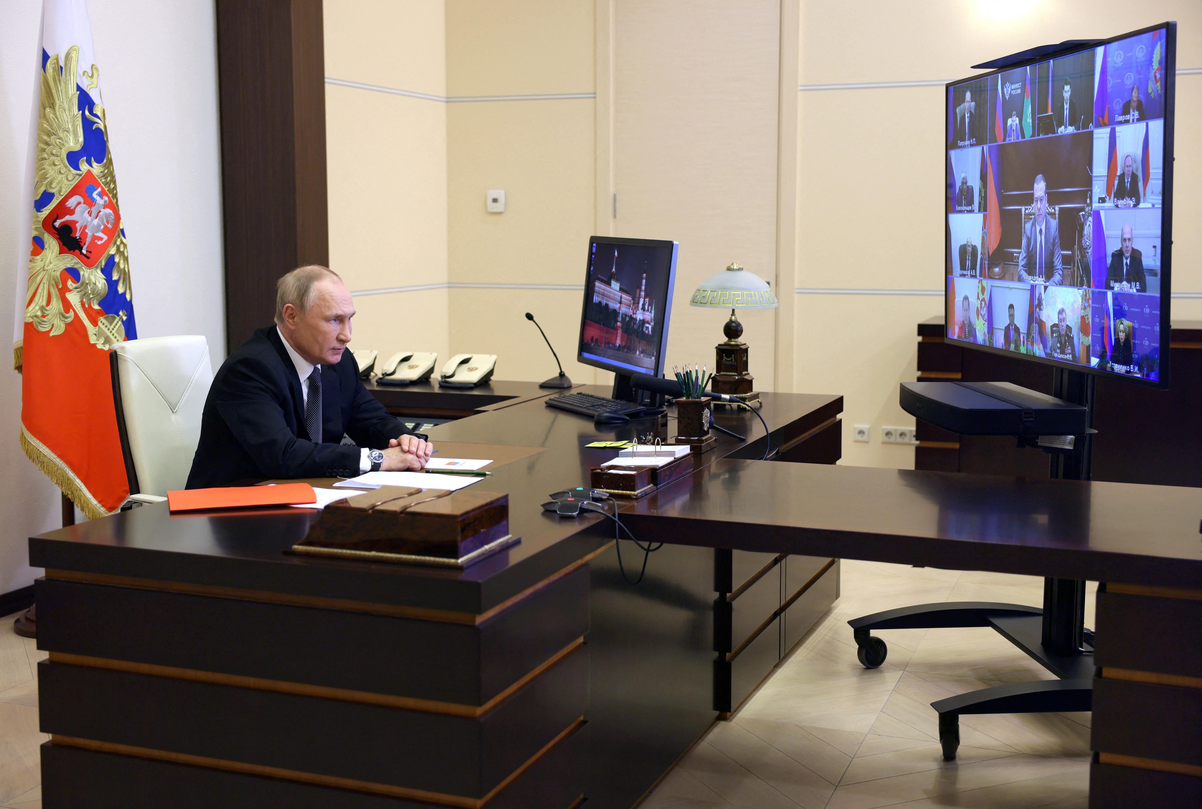 Vladimir Putin sits at a large wooden desk in front of a video screen.