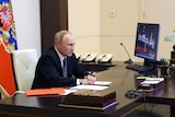 Vladimir Putin sits at a large wooden desk in front of a video screen.