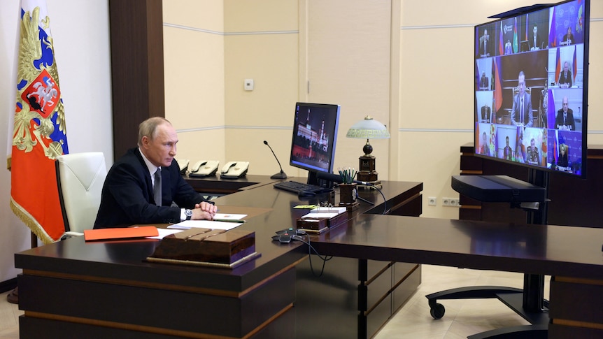 Vladimir Putin sits at a large wooden desk in front of a video screen.