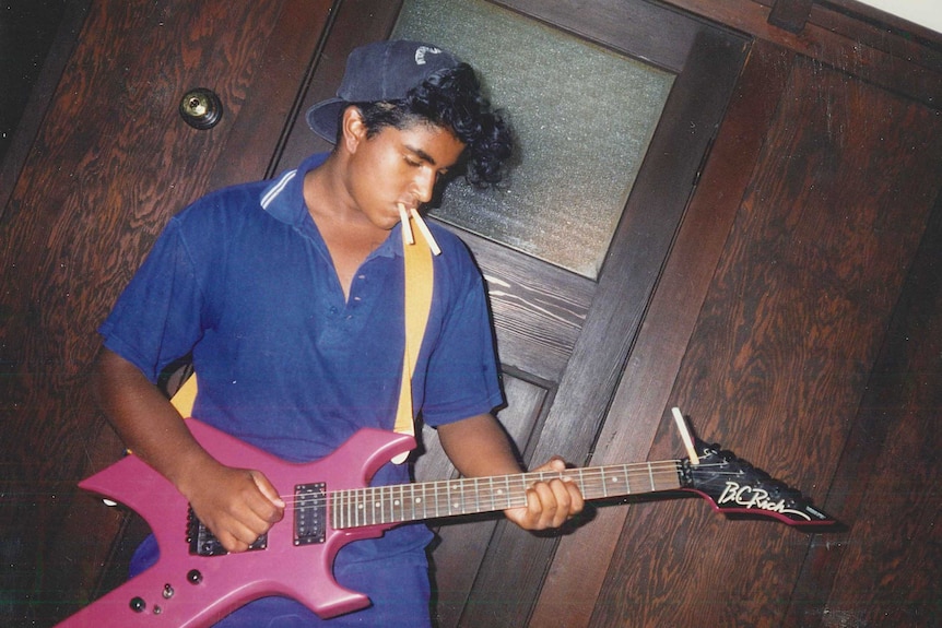 A man with his hat backwards playing a guitar with two cigarettes in his mouth