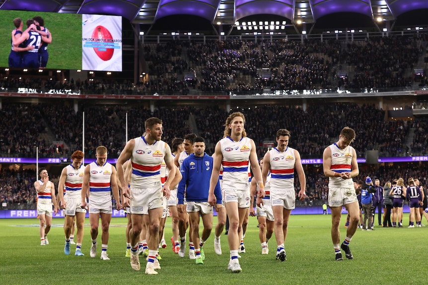 A group of dejected AFL players trudge off the ground after a finals loss, while winners' celebrations are on the big screen.