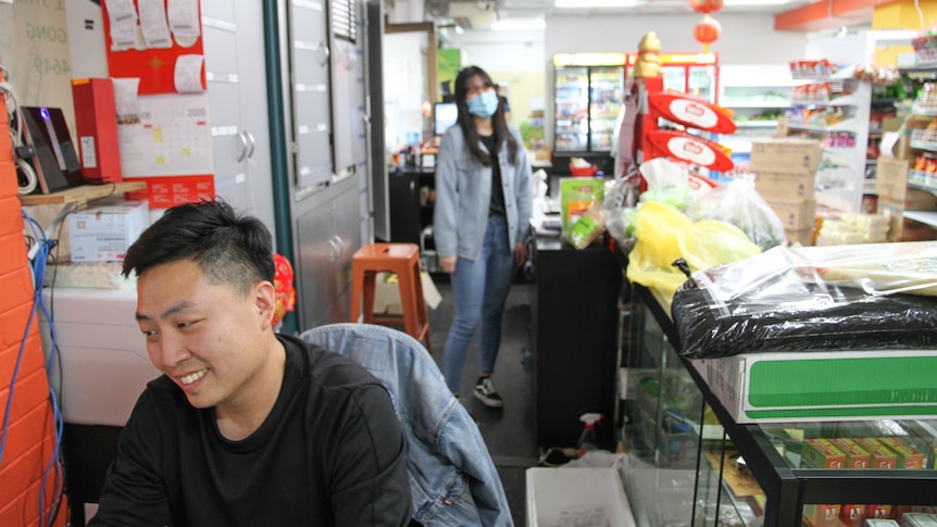 A man sits working whilst a girl wearing a mask serves customers