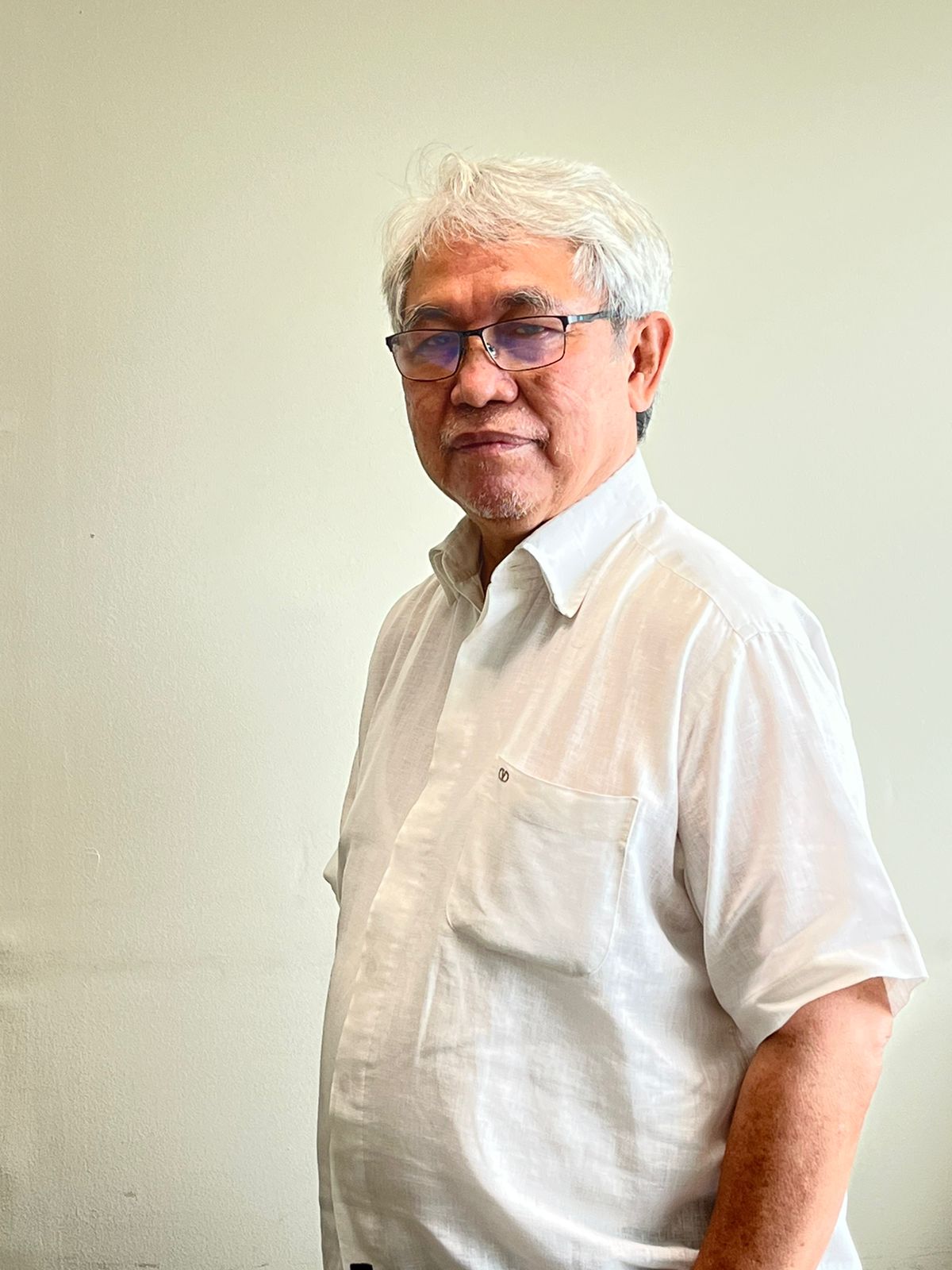 A man around late 60s looking wearing white shirt and glasses looking at the camera.