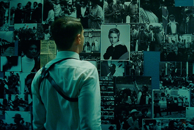 A man in white shirt and suspenders stands facing a large board full of black and white surveillance photos in a dimly lit room.