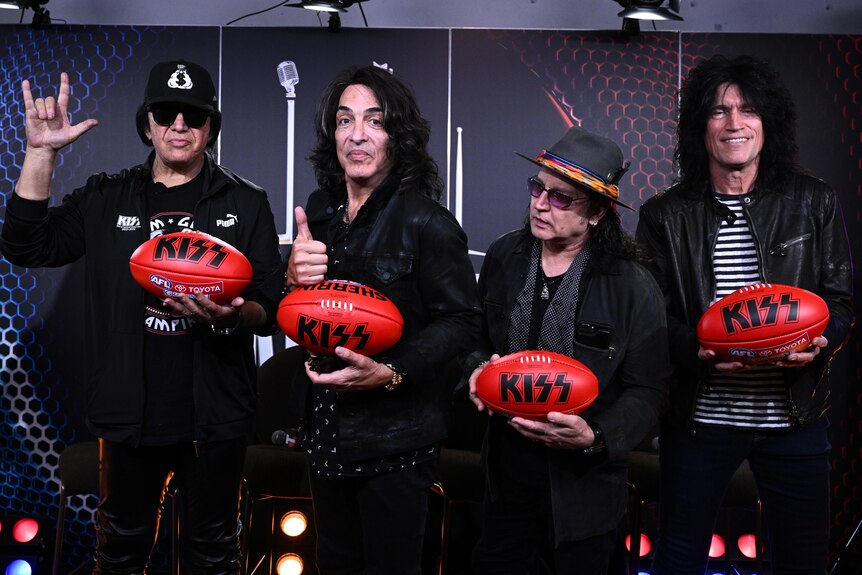 Four members of the band KISS stand giving thumbs up and other gestures as they hold Australian rules footballs.