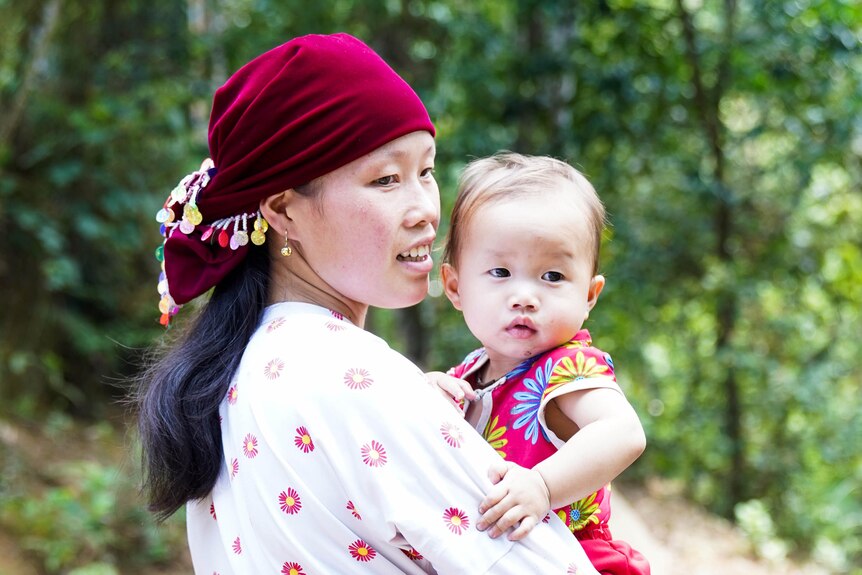 Mai, wearing a maroon traditional headscarf, while holding her baby girl.