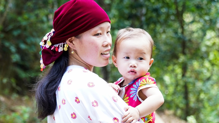 Mai, wearing a maroon traditional headscarf, while holding her baby girl.