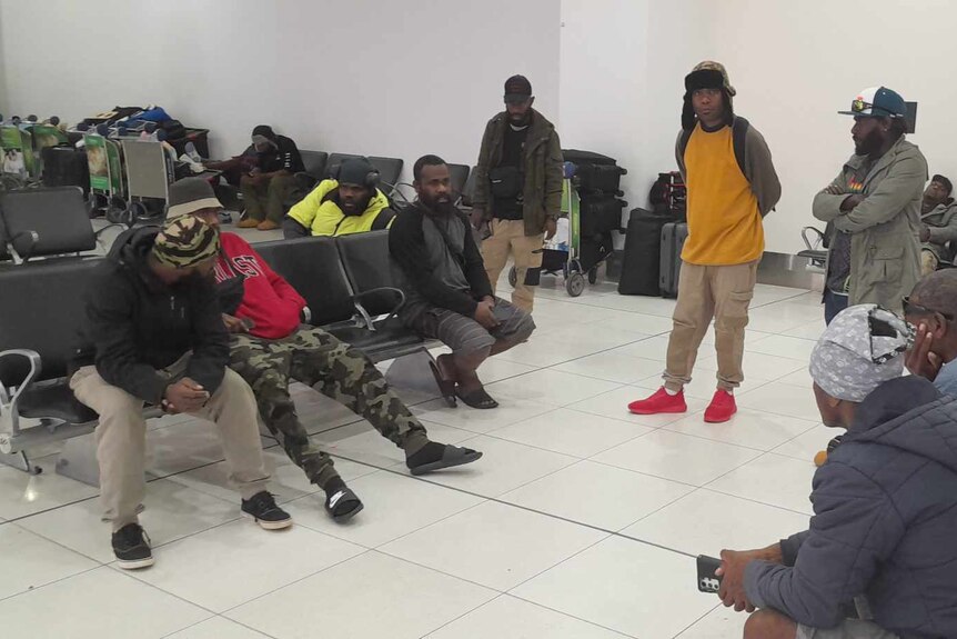 Men sit down and stand waiting at the airport.