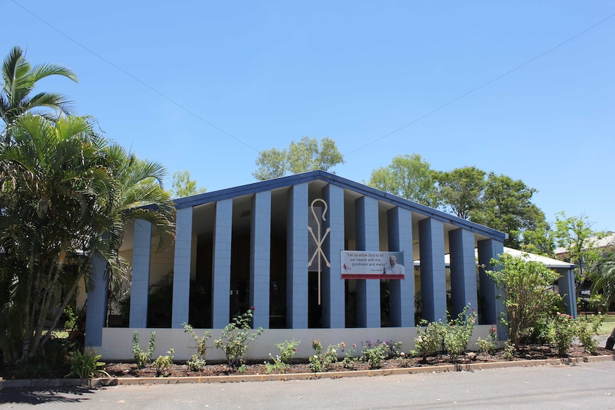 Exterior of a church, with blue-painted columns, a crook-and-cross symbol, and rose bushes growing in front of it.