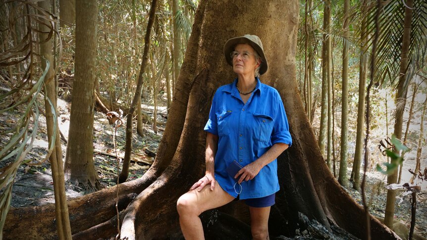 An older woman stands against a tree in a burnt rainforest