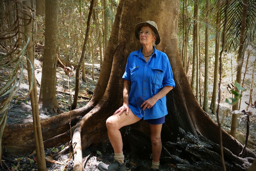 An older woman stands against a tree in a burnt rainforest