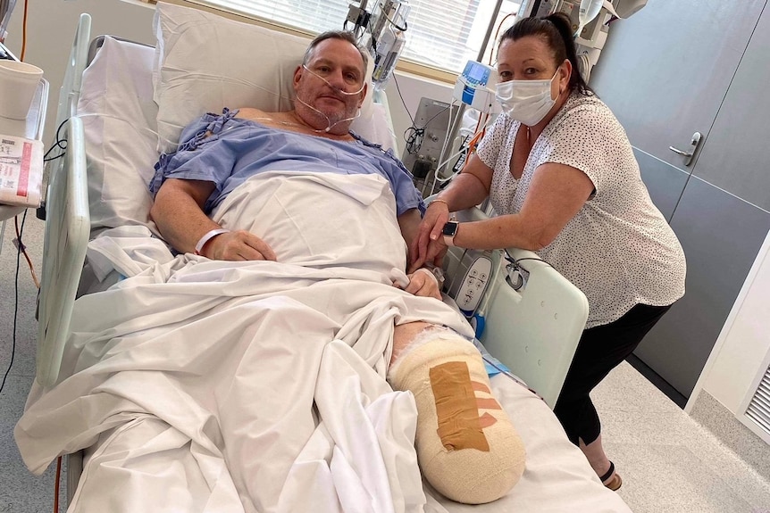 A middle aged man lies in a hospital bed with a bandage covering a stump where his left foot used to be, next to a masked woman