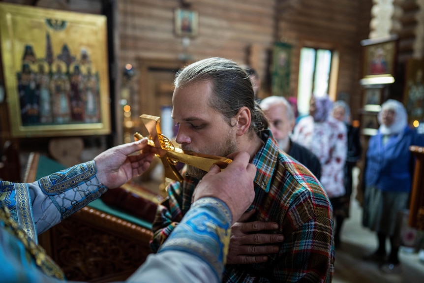 A man with tied-back blond hair kisses a golden cross during a church ceremony.