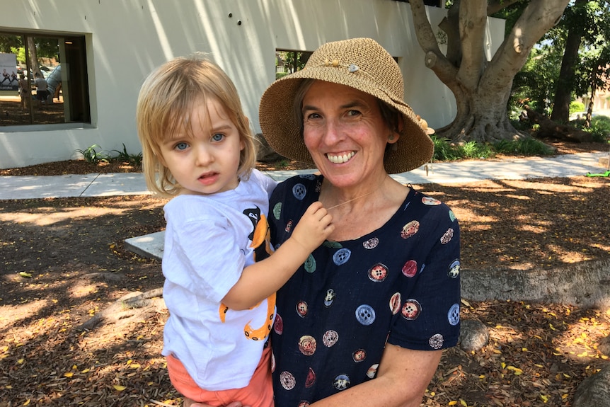 A woman holding a child smiles at the camera while standing in a park.