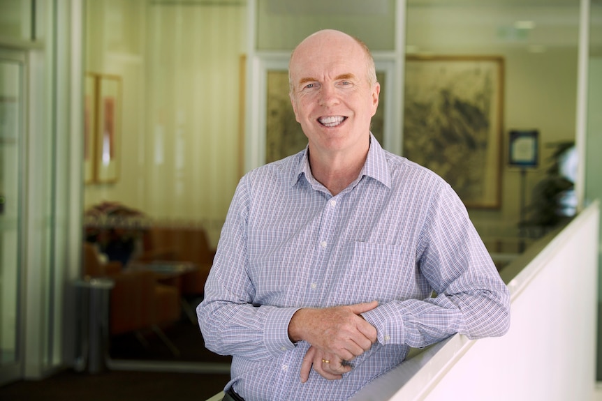 A older bald man in shirt stands smiling in a room. 