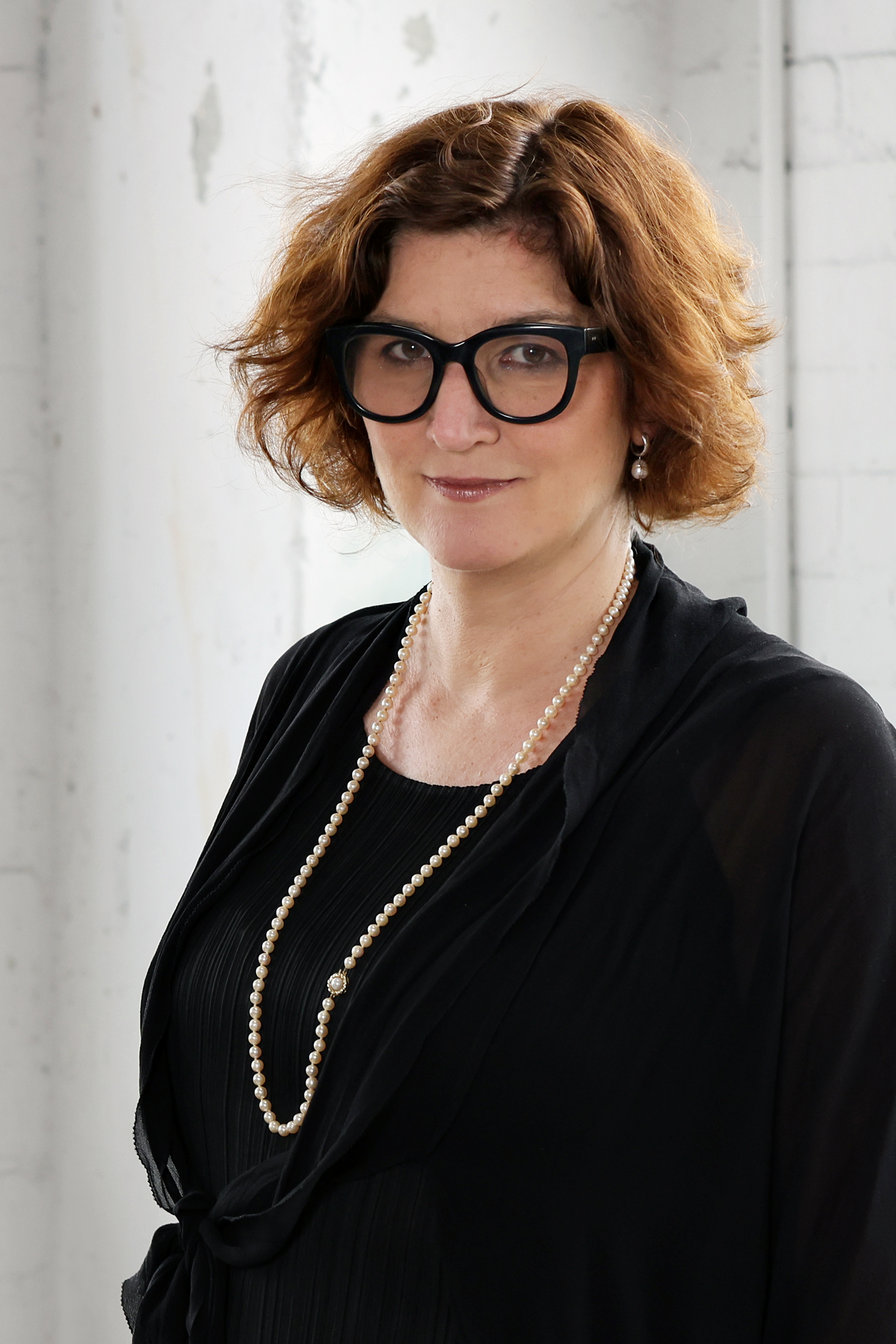 Middle-aged white woman with short, wavy auburn hair wears striking black glasses, a black blazer and white pearls.