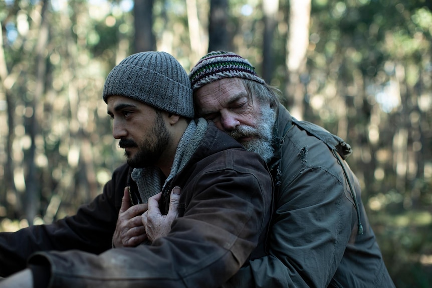 A film still of Phoenix Raei and Hugo Weaving. Weaving has his arms around Raei's middle, hugging him.