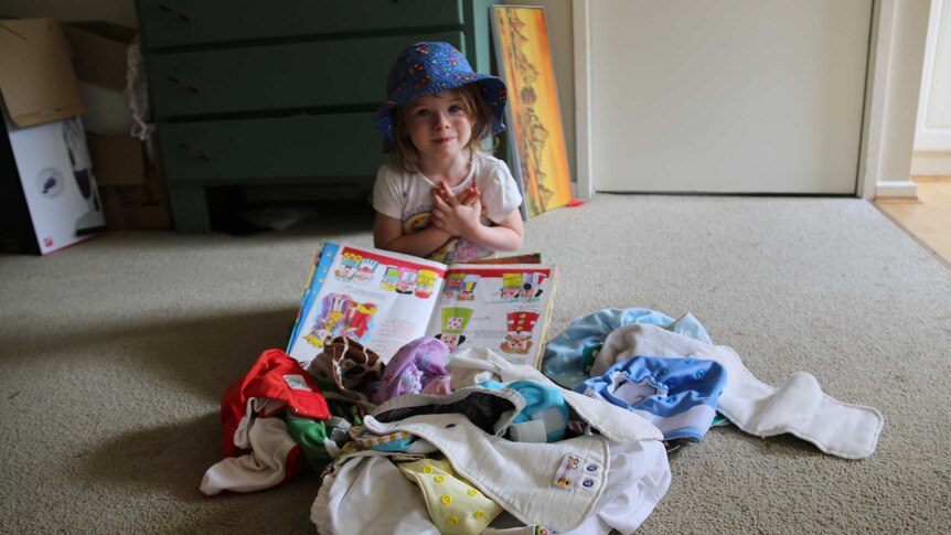 A little girl sits on the floor near a pile of colourful cloth nappies, holding a picture book.