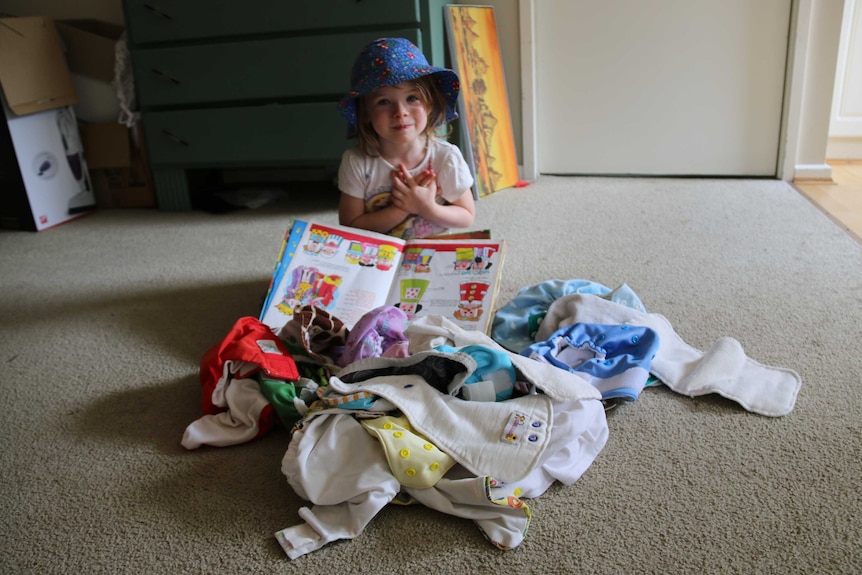 A little girl sits on the floor near a pile of colourful cloth nappies, holding a picture book.