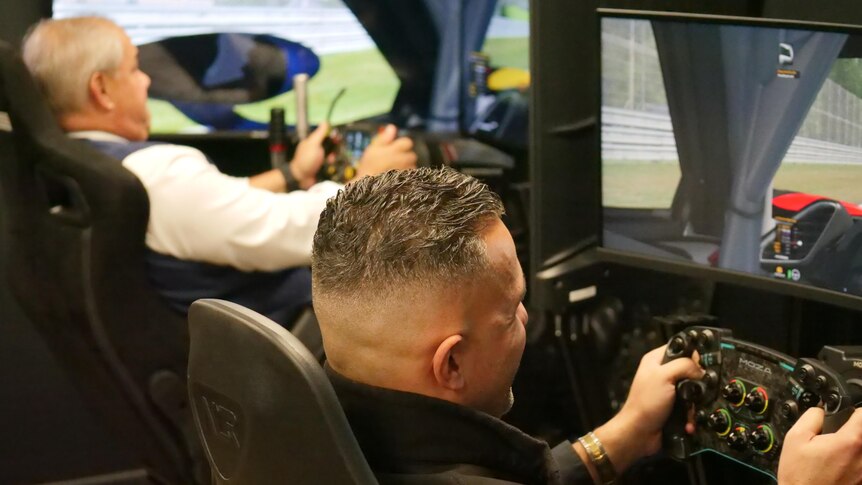 two men driving a car simulator, one wearing white and the other wearing black
