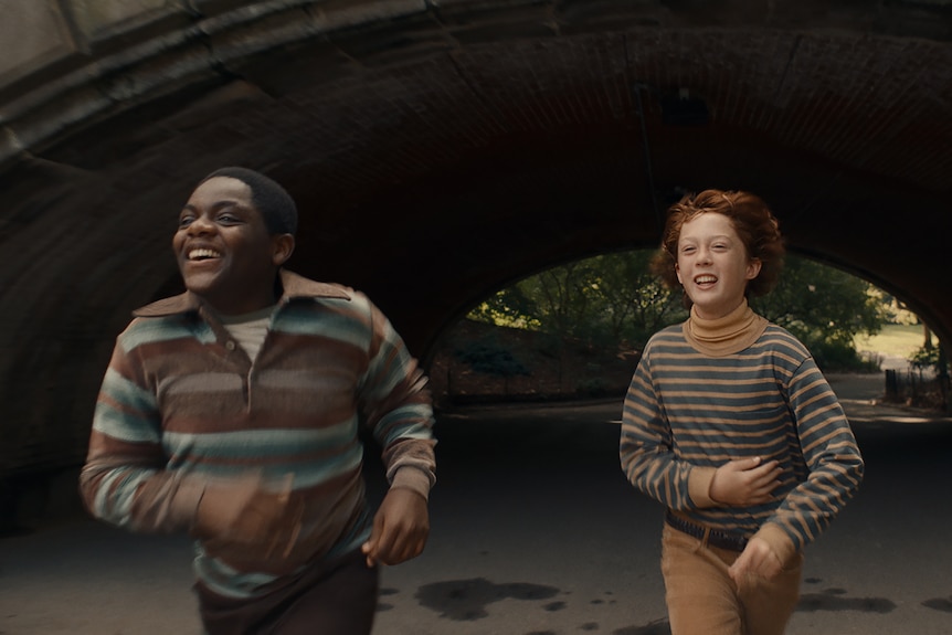 Young Black boy in brown and blue striped shirt runs laughing through tunnel beside white boy in navy and orange striped top.