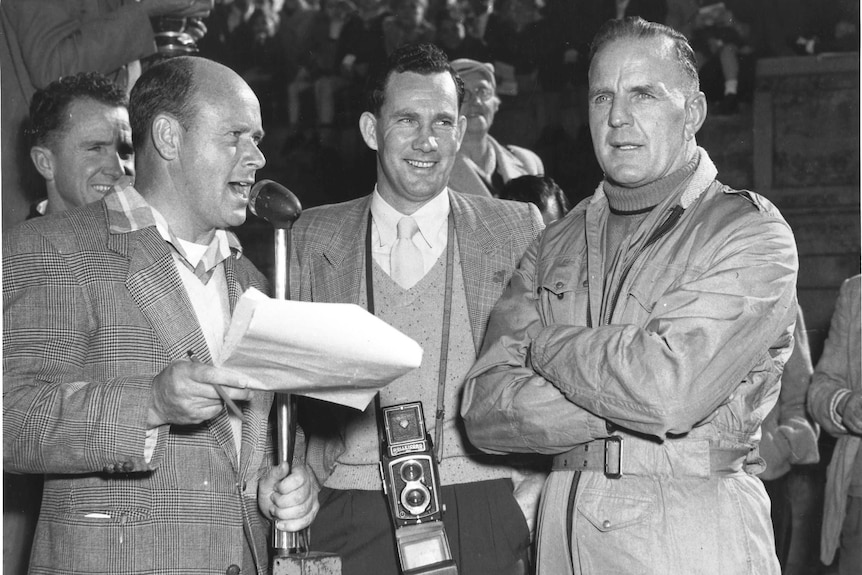 Announcer with microphone, man with a camera, and Jack Murray in a racing jumpsuit.