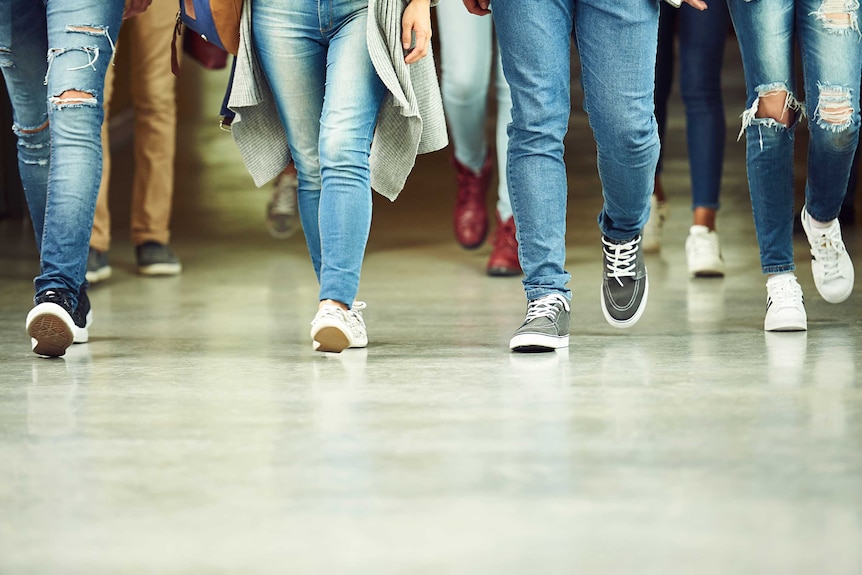 From the waist down, four people in blue jeans and sneakers walk down a hallway. More feet are visible behind them.