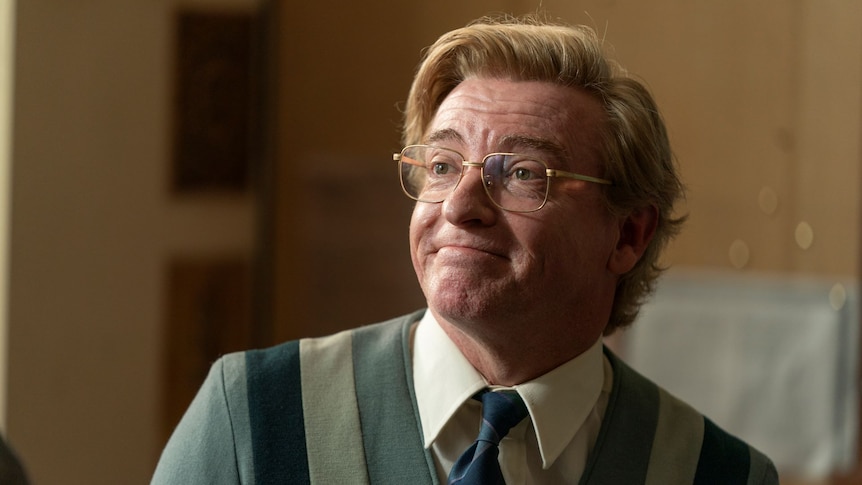 Rhys Darby is pictured in character wearing a daggy cardigan and glasses with a combover. 