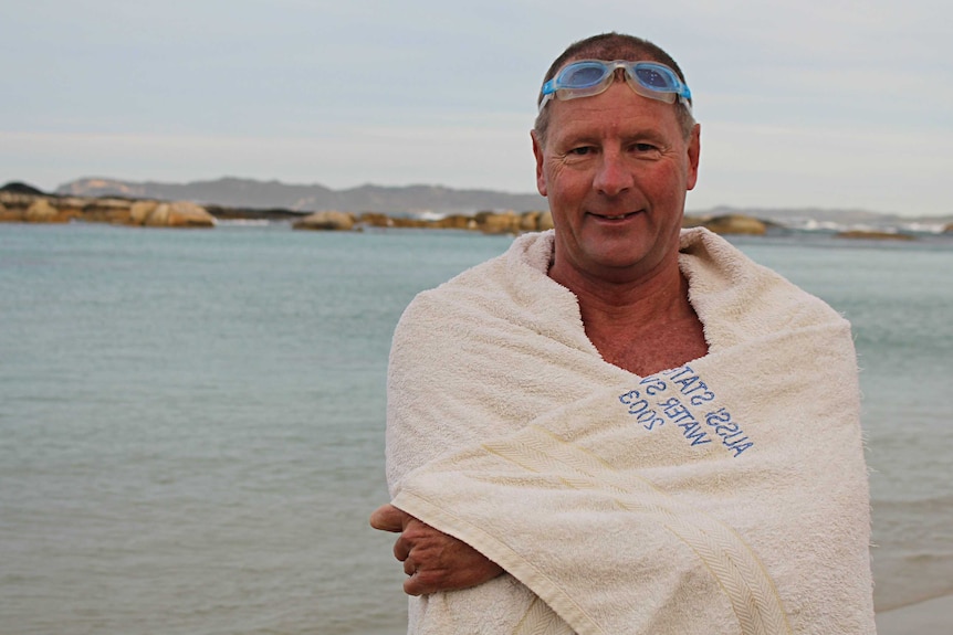 A man wrapped in a white towel with goggles on his head stands on a beach
