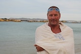 A man wrapped in a white towel with goggles on his head stands on a beach