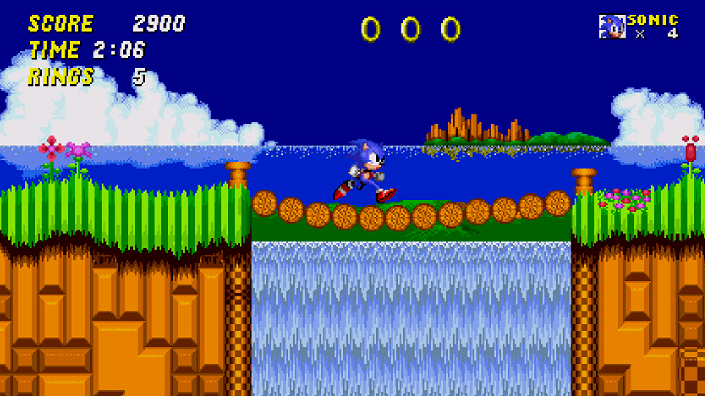 In a scene from a 2D video game a blue hedgehog-like character runs across bridge towards lush grass in location near the ocean.