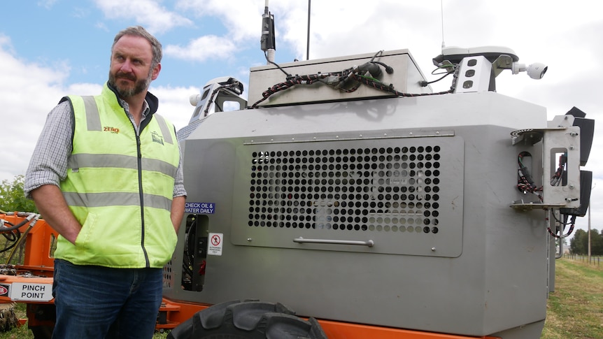 A serious man in hi-vis jacket, looking away, standing on the left next to a grey metal unit, which is a robotic tractor.