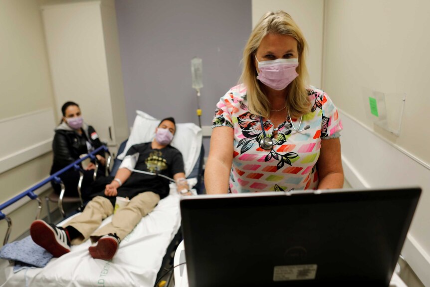 A nurse in a face mask looks at a computer while a man in a bed lies behind her