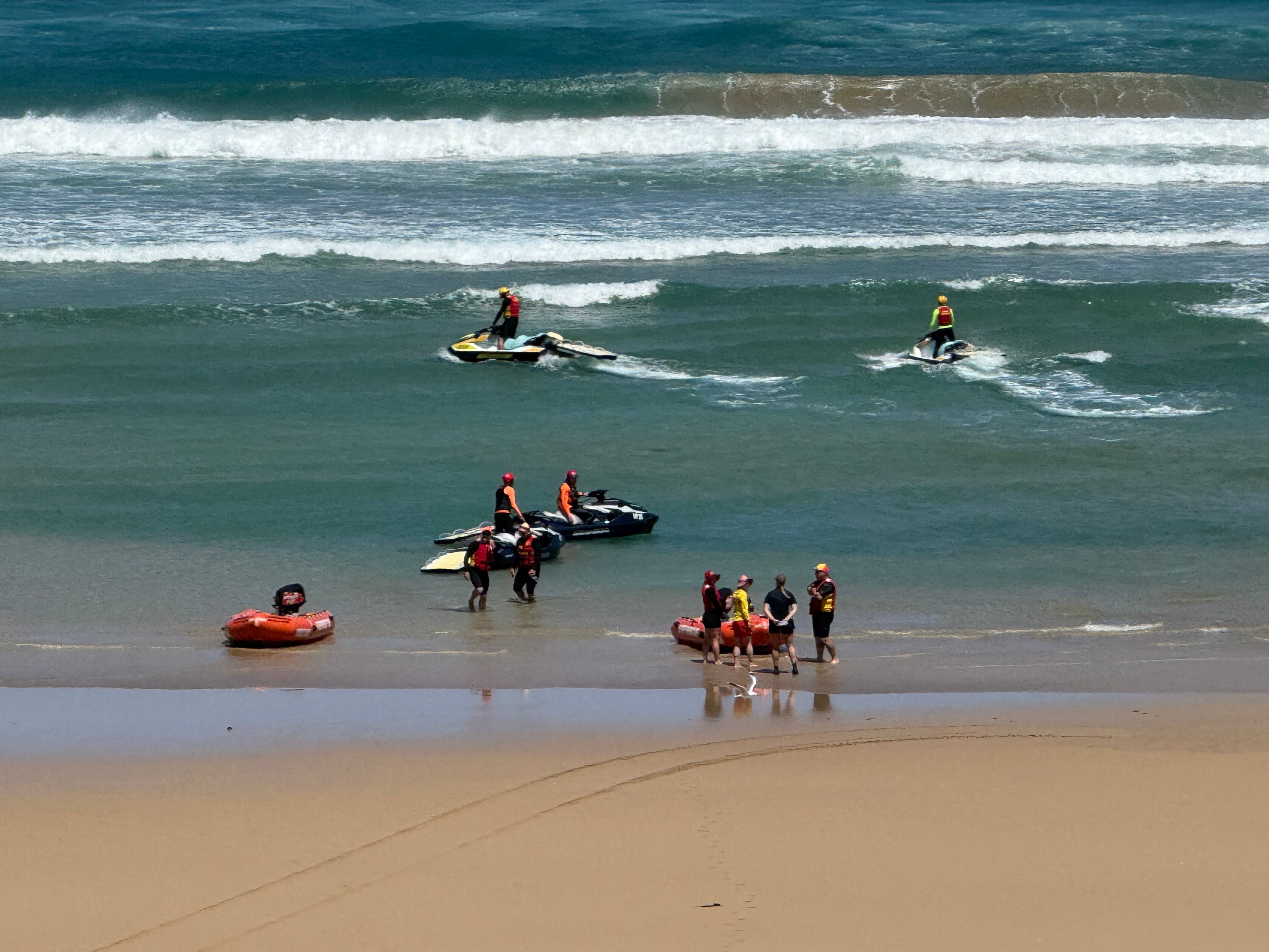 Una imagen de una playa con grupos de búsqueda en inflables naranjas y motos de agua en el agua.