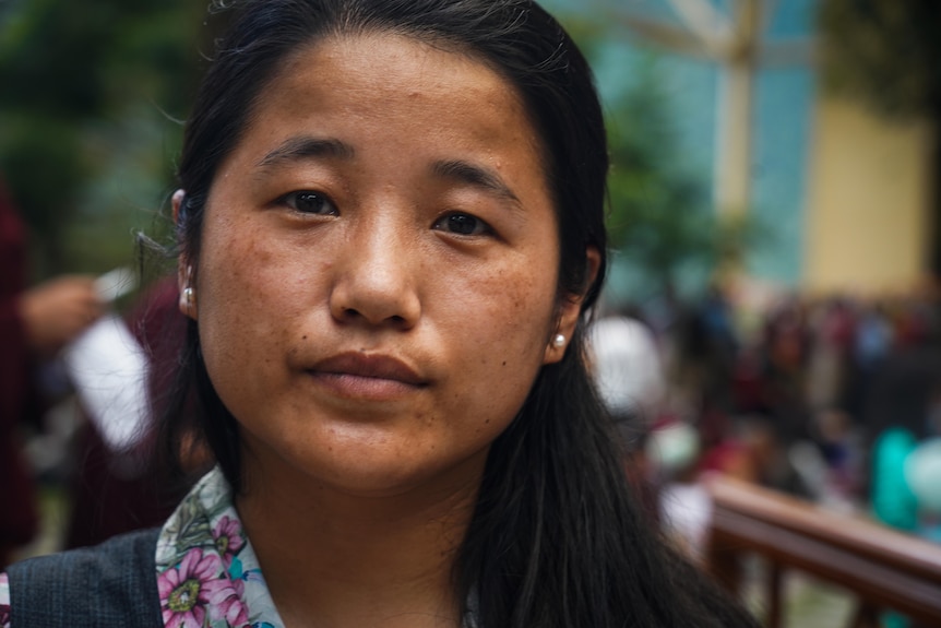 A woman in close-up looks into the camera with serious expression on her face. Her floral collar and pearl earrings are visible