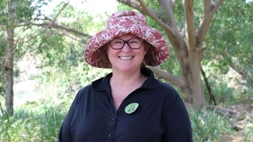 A woman wearing a red and white hat and glasses smiles broadly standing under trees