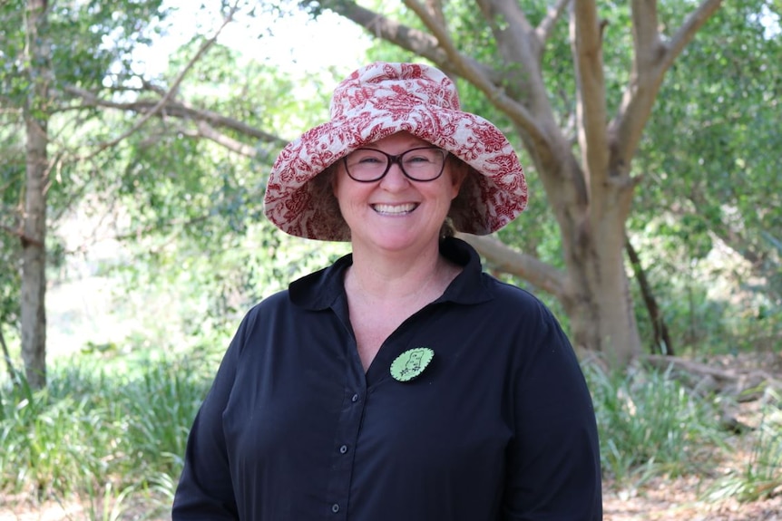 A woman wearing a red and white hat and glasses smiles broadly standing under trees