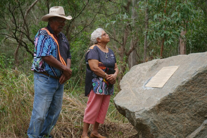 Two Indigenous people stand beside a plaque on a boulder with bush and trees surrounding them.