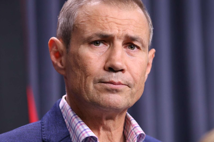 A close up of Roger Cook's face with a blue curtain background, looking concerned.