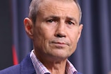 A close up of Roger Cook's face with a blue curtain background, looking concerned.
